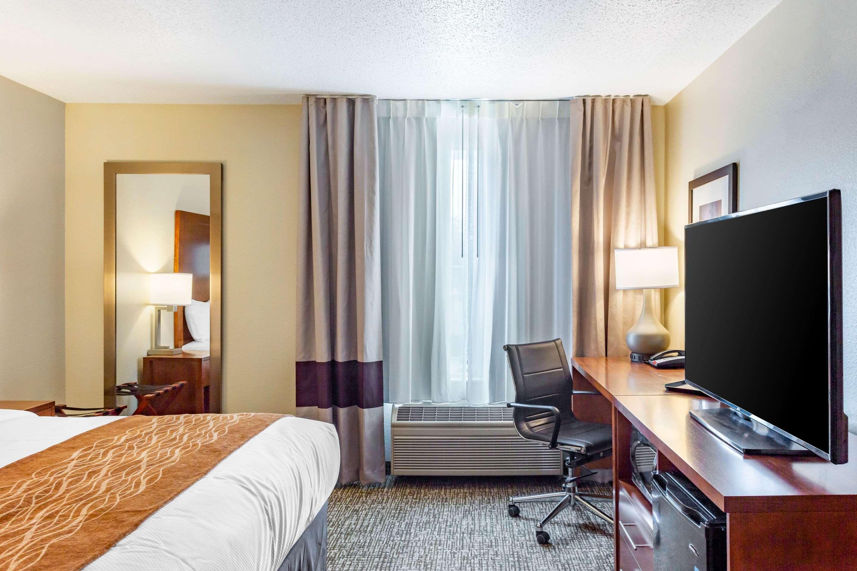A hotel room with a bed, a desk with a television, a chair, and a large window with curtains.