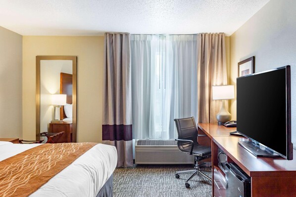 A hotel room with a bed, a desk with a television, a chair, and a large window with curtains. - Comfort Inn (DeKalb)