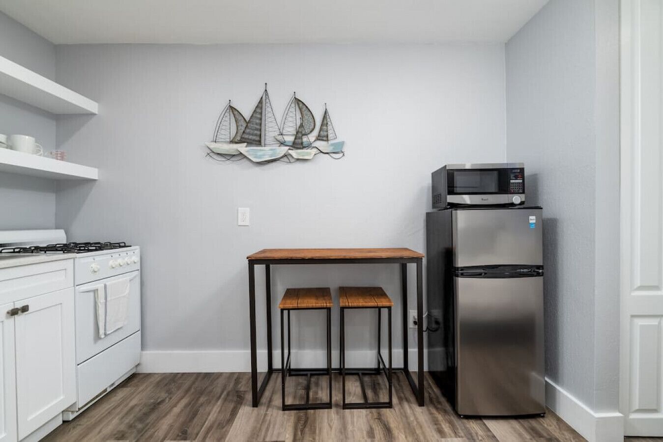 Una cocina compacta con estufa blanca, refrigerador de acero inoxidable, mesa de madera con dos sillas y un adorno náutico de barco en la pared.