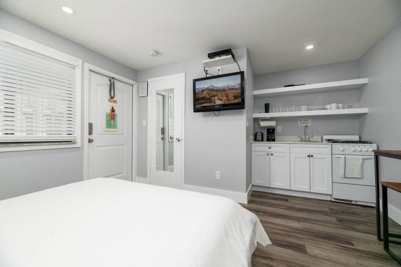 Un dormitorio moderno con una cama grande, un televisor montado en la pared y una cocina con armarios blancos y una estufa.