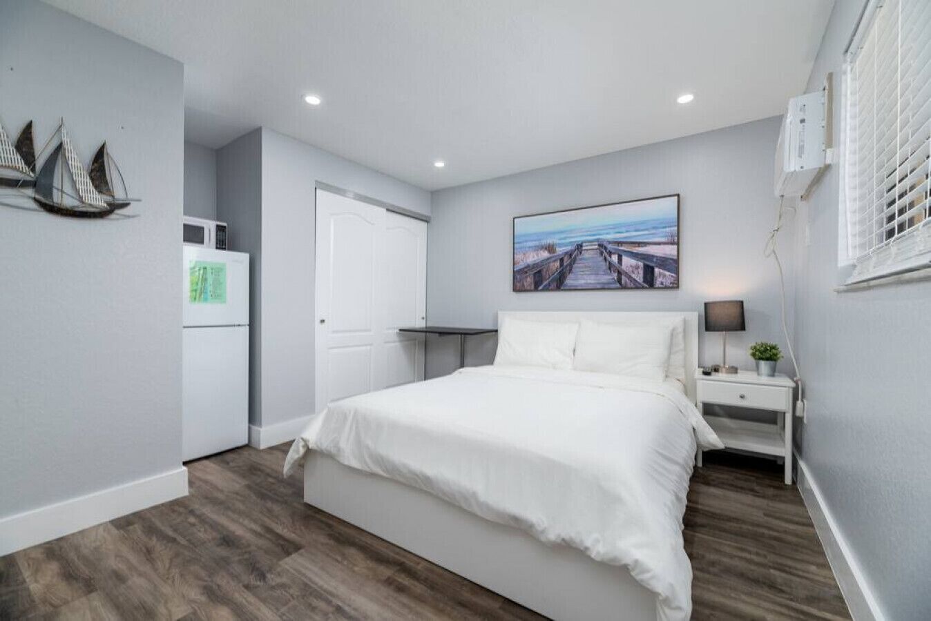 A bedroom with a bed, a nightstand, a lamp, a painting, and a window with blinds.