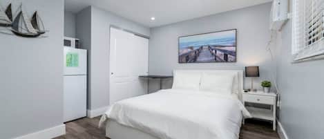 A bedroom with a bed, a nightstand, a lamp, a painting, and a window with blinds.