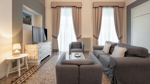 Moderni hotellihuone, jossa on sohva, nojatuoli ja pieni pöytä. Seinällä on taulu ja ikkunoiden alla patteri.