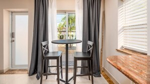 Une petite pièce avec une table ronde et deux chaises, une fenêtre avec des stores et une porte.