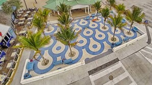 A beachfront hotel with a central courtyard featuring palm trees and a patterned floor, a pier extending into the ocean, and a beach area with umbrellas and lounge chairs. - Ocean Flats #3 - Lauderdale-by-the-Sea (Lauderdale-by-the-Sea)
