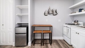 A compact kitchen with white cabinetry, a stainless steel refrigerator, a wooden table with two stools, and a decorative sailboat wall art piece. - Shell on the Beach #103 (Hollywood)