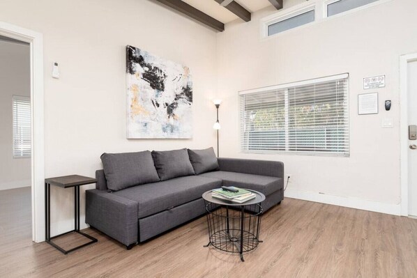 A modern living room with a grey sofa, a small round table with books, a large abstract painting on the wall, and a window with blinds.