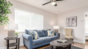 Ein Wohnzimmer mit einem blauen Sofa, einem runden Couchtisch, einem Beistelltisch mit einer Lampe, einer Zimmerpflanze und einem Deckenventilator.