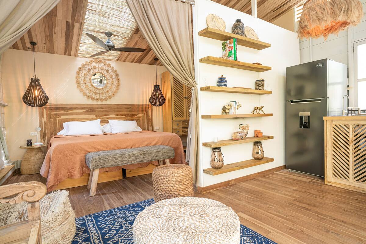 Une chambre avec un lit en bois, un ventilateur de plafond, une armoire intégrée et une kitchenette équipée d’un réfrigérateur.