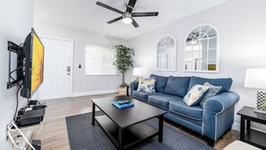 Un salon moderne avec un canapé bleu, une table basse, un téléviseur fixé au mur et un ventilateur de plafond.