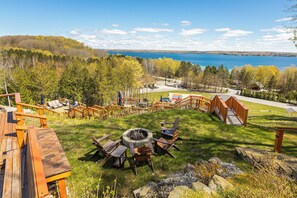 A deck with outdoor furniture, a fire pit, and a view of a lake. - Luxury Torch Lake Vacation Home – Private Dock, Pickleball, Game Room, Theatre (Kewadin)