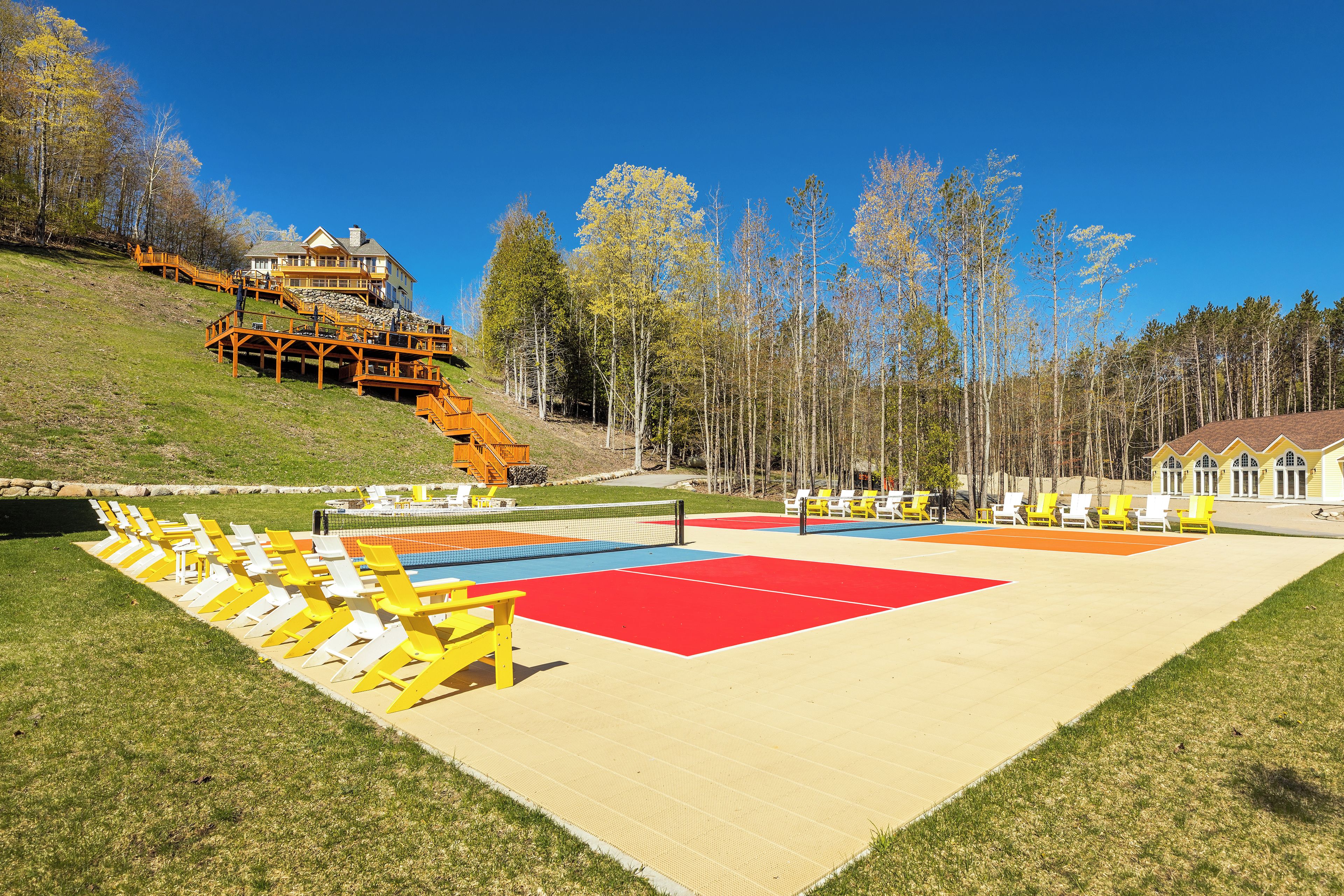 A beach volleyball court with yellow and white chairs, a red and blue playing area, and a wooden staircase leading to a building.
