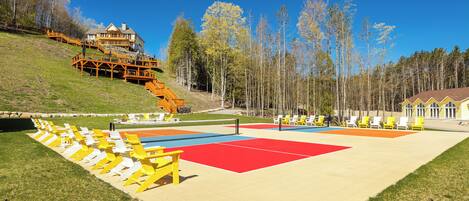 A beach volleyball court with yellow and white chairs, a red and blue playing area, and a wooden staircase leading to a building.
