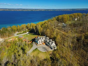 A large house with a driveway and a yard, surrounded by trees and a lake. - Luxury Torch Lake Vacation Home – Private Dock, Pickleball, Game Room, Theatre (Kewadin)