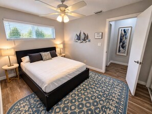 A bedroom with a bed, bedside lamps, a ceiling fan, a window with blinds, a patterned rug, and a door leading to another room.