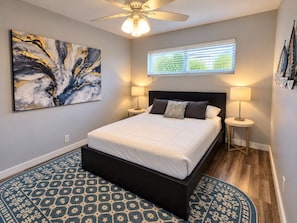 A bedroom with a bed, two bedside lamps, a ceiling fan, a window with blinds, and a large abstract painting on the wall. - The Palms @ Middle River Terrace #3 (Fort Lauderdale)