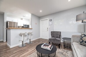 A modern kitchen with white cabinets, a microwave, and a stainless steel refrigerator. There is a dining area with a grey upholstered chair and a small round table. A sofa with patterned cushions is visible in the living room area.