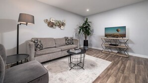 A modern living room with a grey sofa, a coffee table, a floor lamp, a TV on a wooden stand, and a decorative wall piece. - The Palms @ Middle River Terrace #3 (Fort Lauderdale)