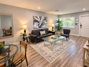 A modern living room with a glass coffee table, a black leather sofa, a dining area with a glass table, and a large abstract painting on the wall. - The Palms @ Middle River Terrace #2 (Fort Lauderdale)