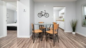 A modern apartment with a dining area featuring a round glass table and four wooden chairs. A bicycle is mounted on the wall above the table. The apartment has a wooden floor, white walls, and a kitchen with white cabinets and a countertop.