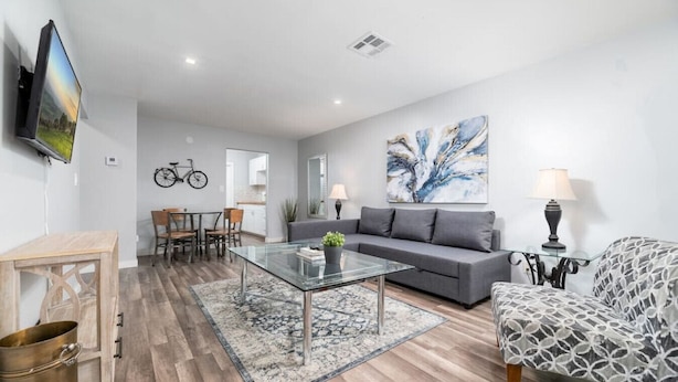 A modern living room with a glass coffee table, grey sofa, patterned armchair, and a dining area with chairs and a table.