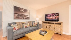 Uma sala de estar com um sofá cinzento, uma mesa de centro de madeira, uma TV sobre um móvel de madeira e um quadro com tema praiano na parede.