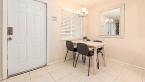 Uma sala de jantar com uma porta branca, uma mesa com quatro cadeiras, uma janela com persianas e um espelho fixado na parede.