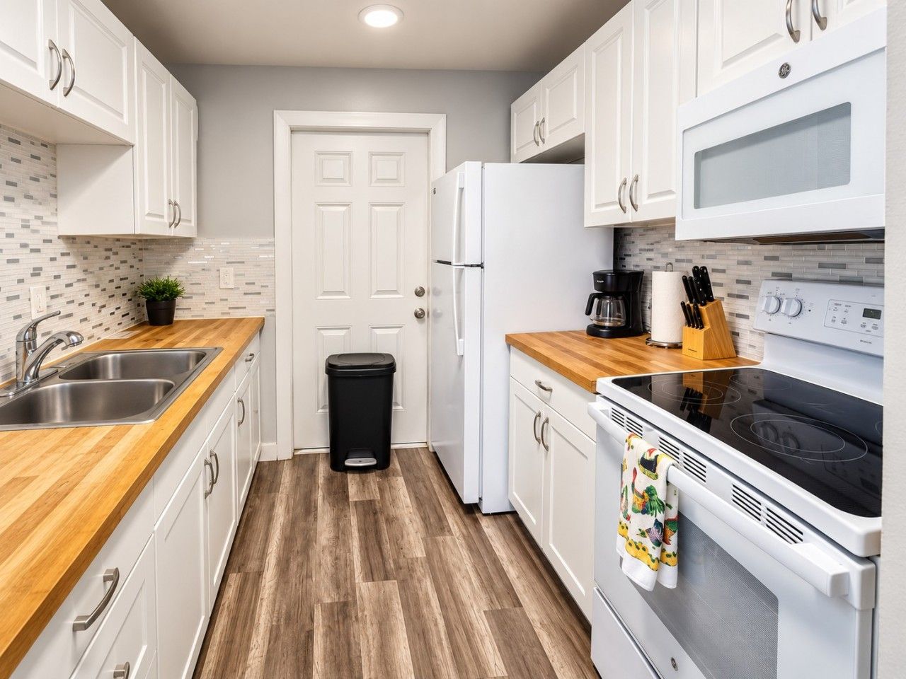 A compact kitchen with white cabinets, a stainless steel sink, a black trash bin, a white refrigerator, a microwave, and an oven with a wooden countertop.