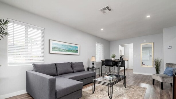 A modern living room with a grey sectional sofa, a glass coffee table, and a dining area with a black chair.
