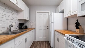 A compact kitchen with white cabinets, a wooden countertop, a stainless steel sink, a coffee maker, a refrigerator, and a dish rack.