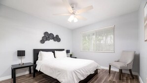 A bedroom with a bed, a chair, a nightstand, a lamp, a ceiling fan, and a window with blinds.