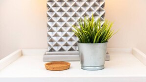 Un secchio di metallo con piante verdi su uno scaffale bianco, un sottobicchiere di sughero e un pannello decorativo geometrico.
