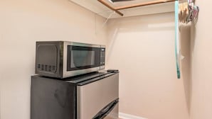 Un angolo cottura compatto con un forno a microonde sopra un frigorifero, un pavimento in legno e un mobiletto a parete con prodotti per la pulizia.