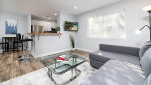 Een moderne woonkamer met een glazen salontafel, een grijze bank en een keuken op de achtergrond.