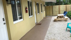 A covered outdoor area with a concrete floor, a wooden table and chairs, a fire extinguisher, and a wooden fence in the background. - The Retreat #1 - Wilton Manors (Wilton Manors)