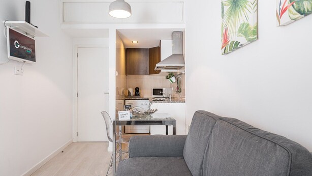 Un salon moderne avec un canapé gris, une table basse en verre et un coin repas comprenant une table et des chaises blanches. On aperçoit une cuisine à l’arrière-plan, avec des meubles en bois et des appareils électroménagers modernes.