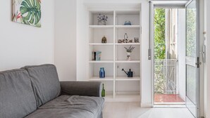Un salon moderne avec un canapé gris, une étagère blanche ornée de divers objets décoratifs et une porte vitrée donnant sur un espace extérieur.