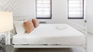 Une chambre d’hôtel moderne avec un lit blanc, deux fenêtres équipées de stores, un mur à motifs et un radiateur blanc.
