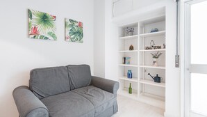 Un canapé gris dans une pièce aux murs blancs, une étagère blanche contenant divers objets et deux œuvres d’art accrochées au mur.