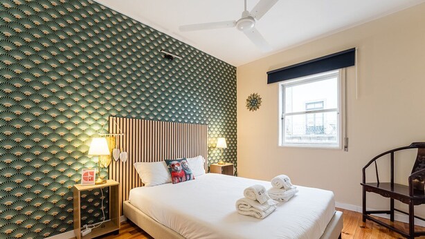 A hotel room with a bed, two bedside tables with lamps, a chair, a window with a view, and a ceiling fan.