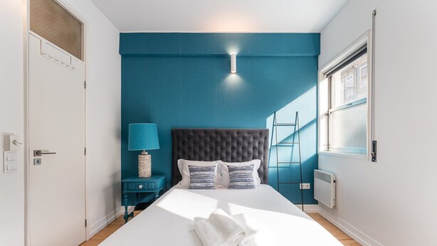 A bedroom with a large bed, a nightstand with a lamp, a window with blinds, and a radiator. - GuestReady Economy - Riber Porto 4R (Porto)