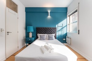 A bedroom with a large bed, a nightstand with a lamp, a window with blinds, and a radiator. - GuestReady Economy - Riber Porto 4R (Porto)