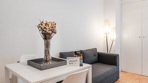 A modern room with a white desk, a grey sofa, a vase with dried flowers, a welcome sign, and a lamp. - GuestReady Economy - Riber Porto 4R (Porto)