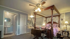 A bedroom with a four-poster bed, a ceiling fan, a bedside table, a nightstand, and a chair.