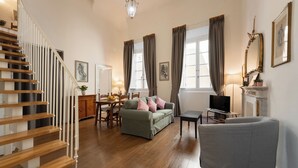 Ein geräumiges Wohnzimmer mit einer Holztreppe, einem grünen Sofa, einem grauen Sessel und einem Essbereich mit Tisch und Stühlen.