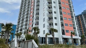 A high-rise residential building with multiple balconies, a wooden walkway, and palm trees. - Oceanfront Studio Suite with breathtaking views (Myrtle Beach)