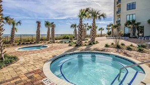 A pool area with palm trees, a building, and a clear sky. - Oceanfront Studio Suite with breathtaking views (Myrtle Beach)