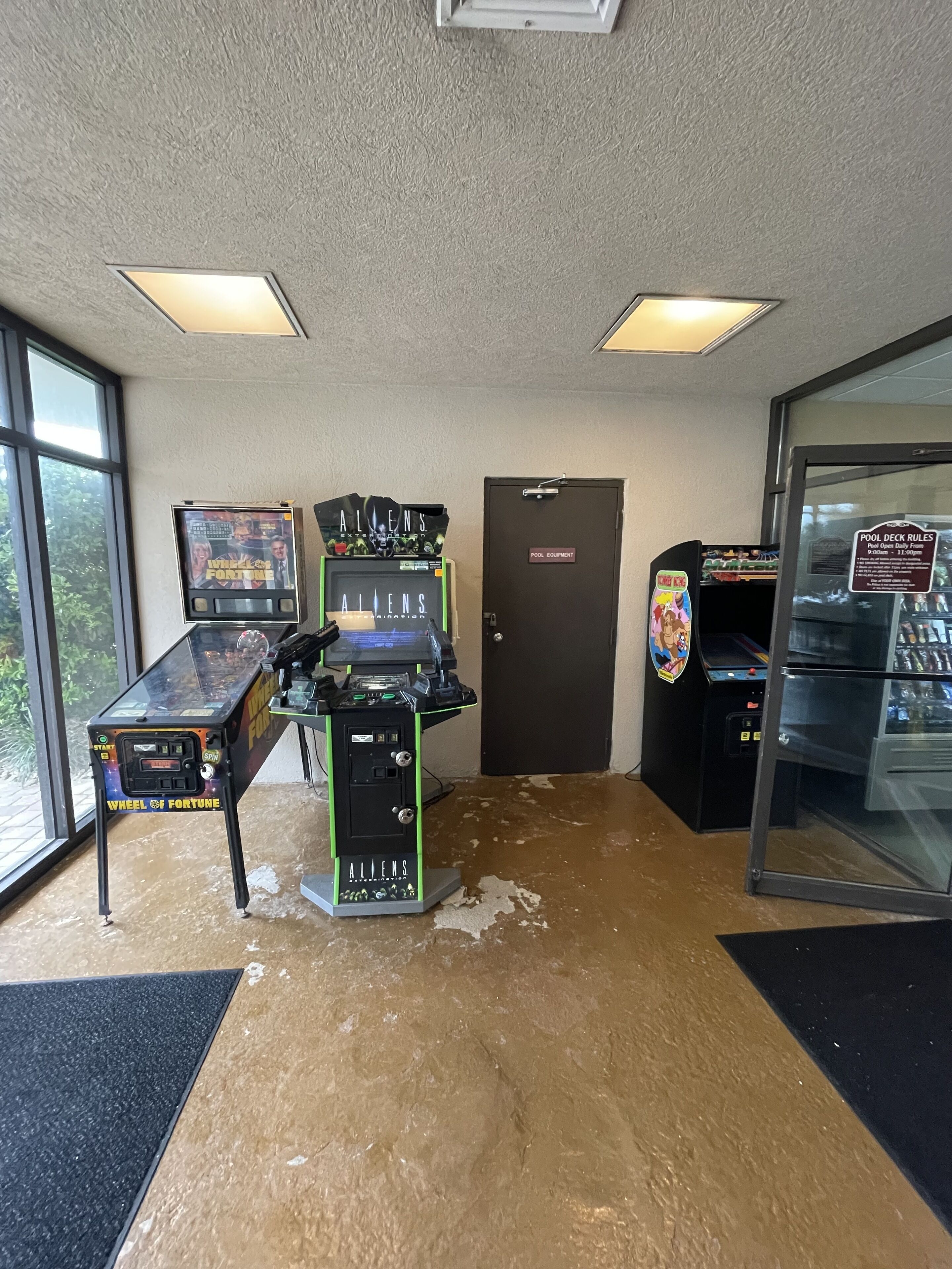 Un espace piscine équipé de jeux d’arcade, d’un panneau indiquant les règles à respecter au bord de la piscine, et d’une porte portant l’inscription « ÉQUIPEMENT DE LA PISCINE ».