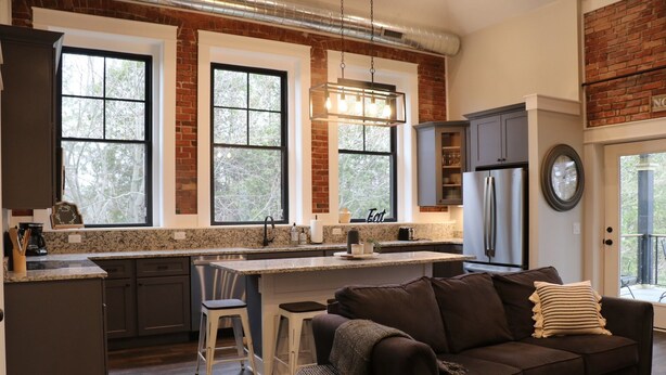 A modern kitchen with a central island, dark cabinetry, and a brick accent wall. - The Sunset View Penthouse Loft (Boyne City)