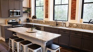 A modern kitchen with granite countertops, a central island with bar stools, stainless steel appliances, and large windows with a view of trees. - The Sunset View Penthouse Loft (Boyne City)